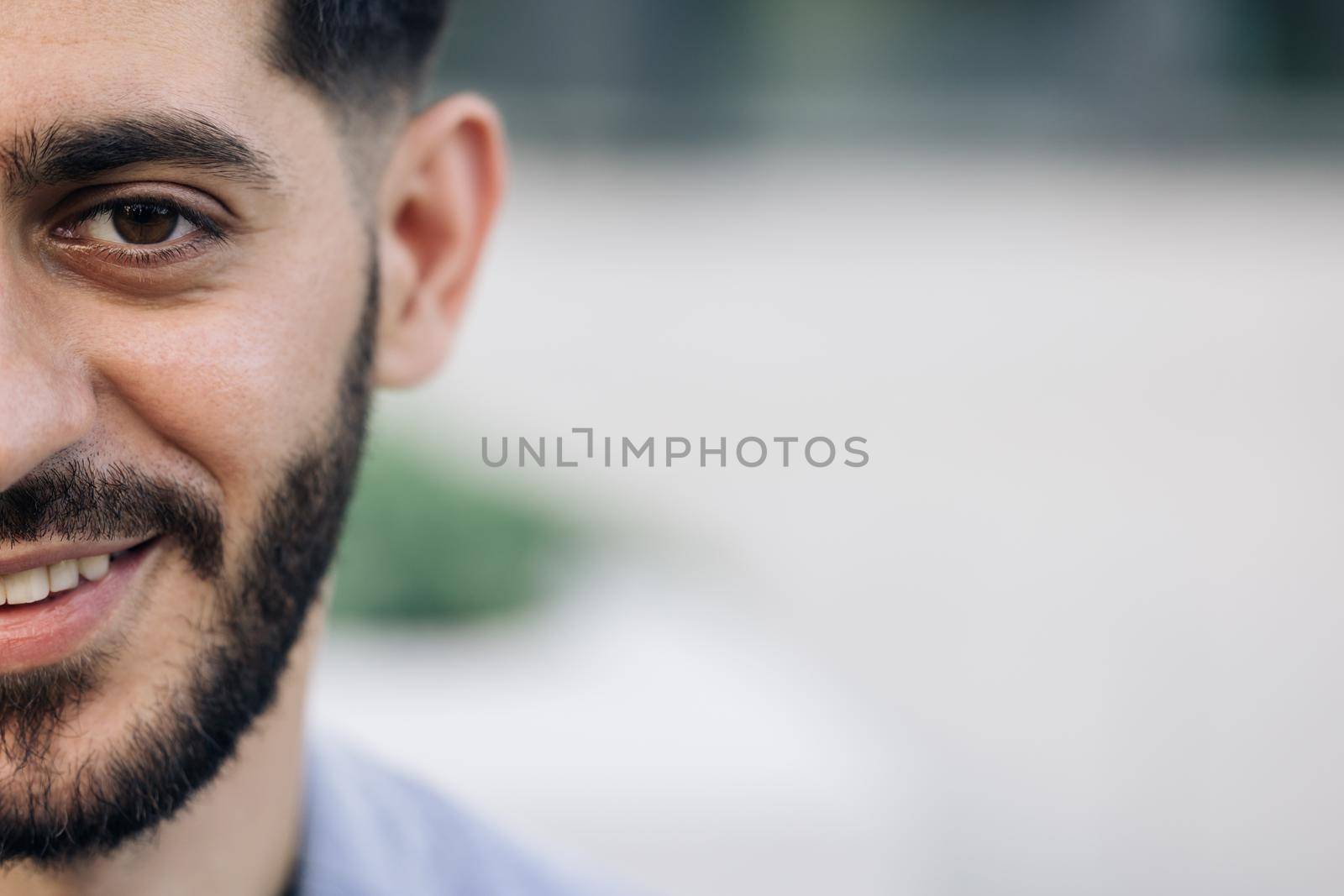 Half face of bearded man with brown eye while standing outdoors. Portrait of male looking to camera and smiling, with space for text. Sensual guy with trendy hairstyle looking into camera.