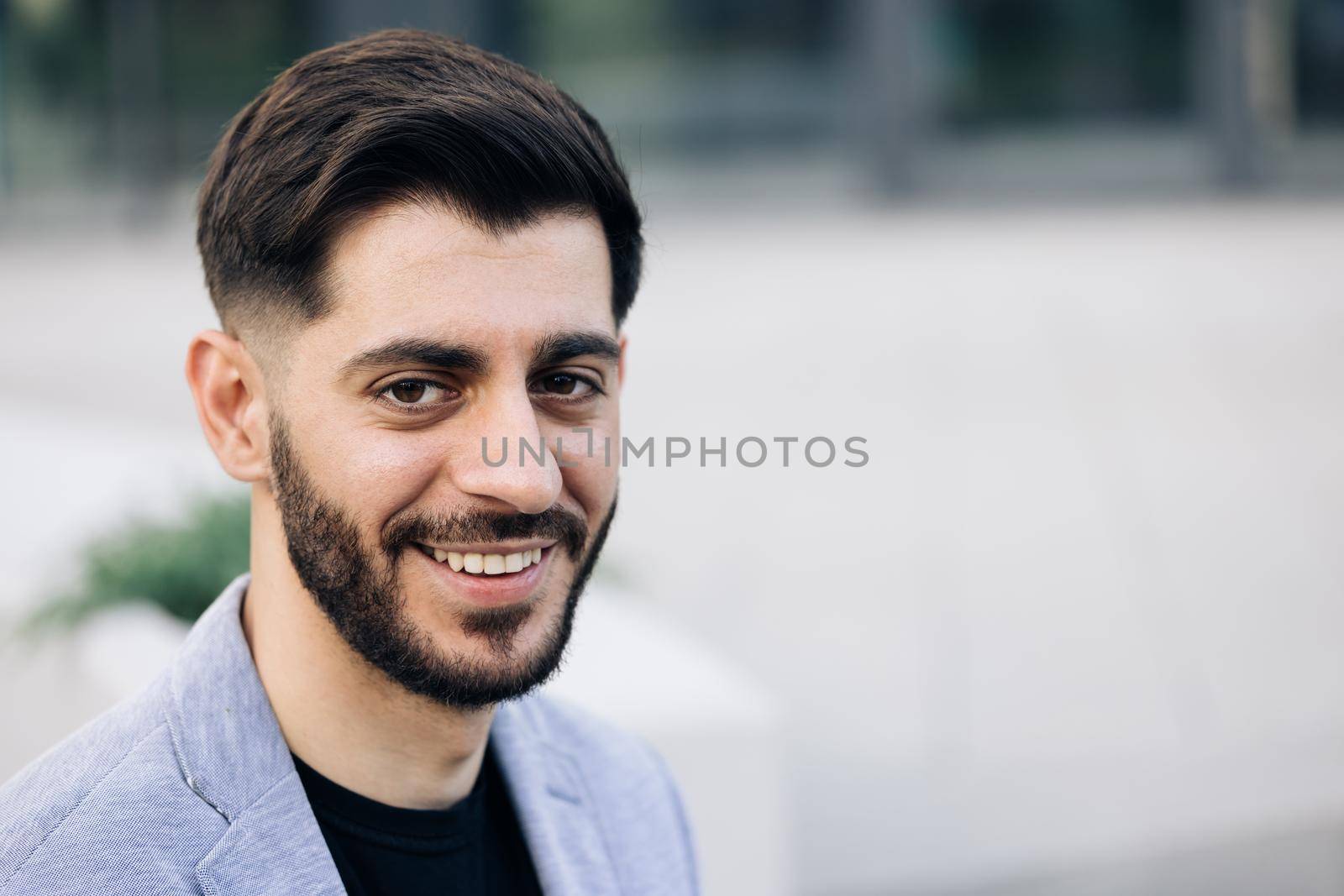 Face portrait of attractive joyful young caucasian bearded man in business formal suit smiling and posing near modern buildings outside. Businessman. Ethnicity.