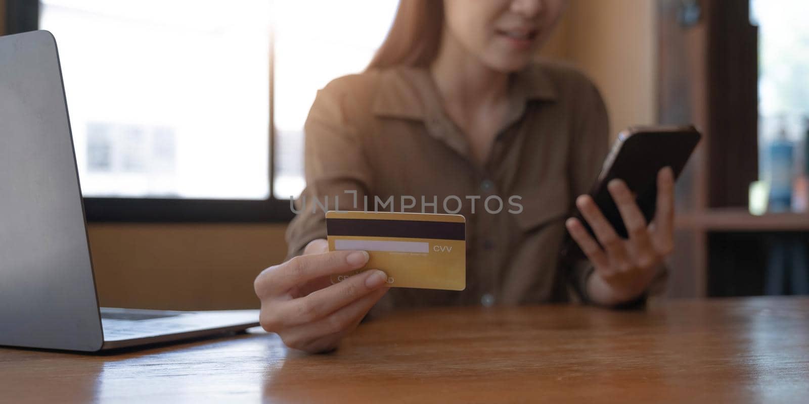 Closeup image of a woman's hand holding credit card and pressing at mobile phone on wooden table in office.
