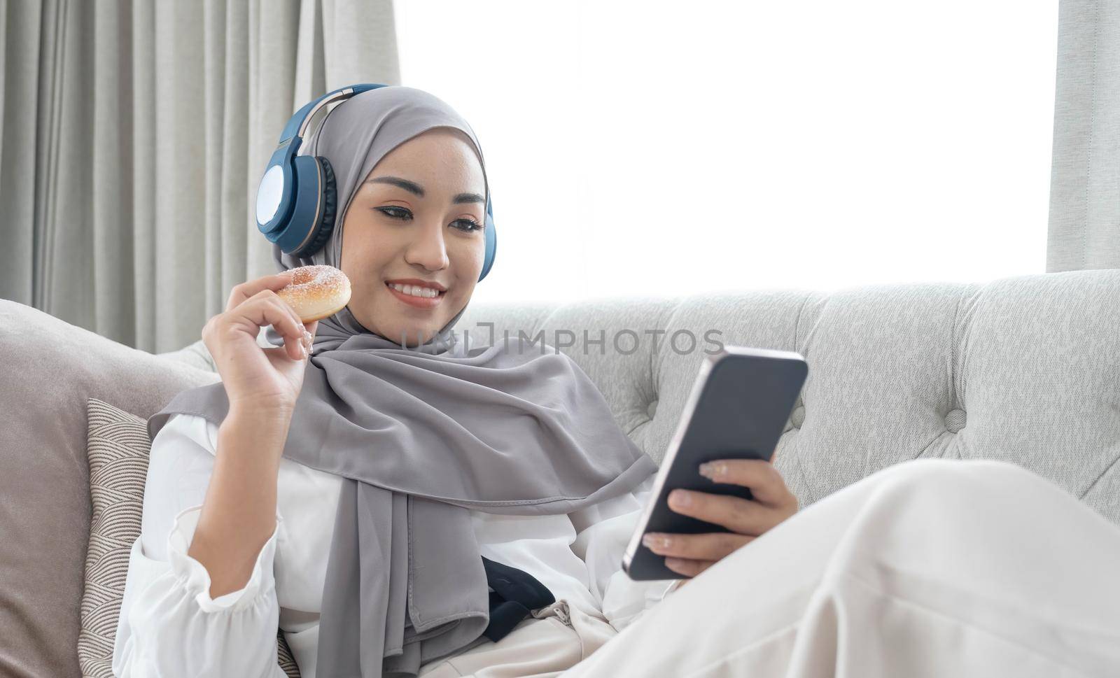 Attractive young Asian Muslim woman wearing hijab and headphones, watching movie on smartphone and eating doughnuts on sofa. by wichayada