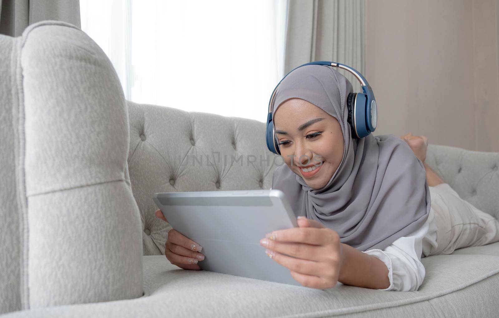 Relaxed and happy asian Muslim woman with hijab laying on sofa, listening music on headphones and using digital tablet. by wichayada