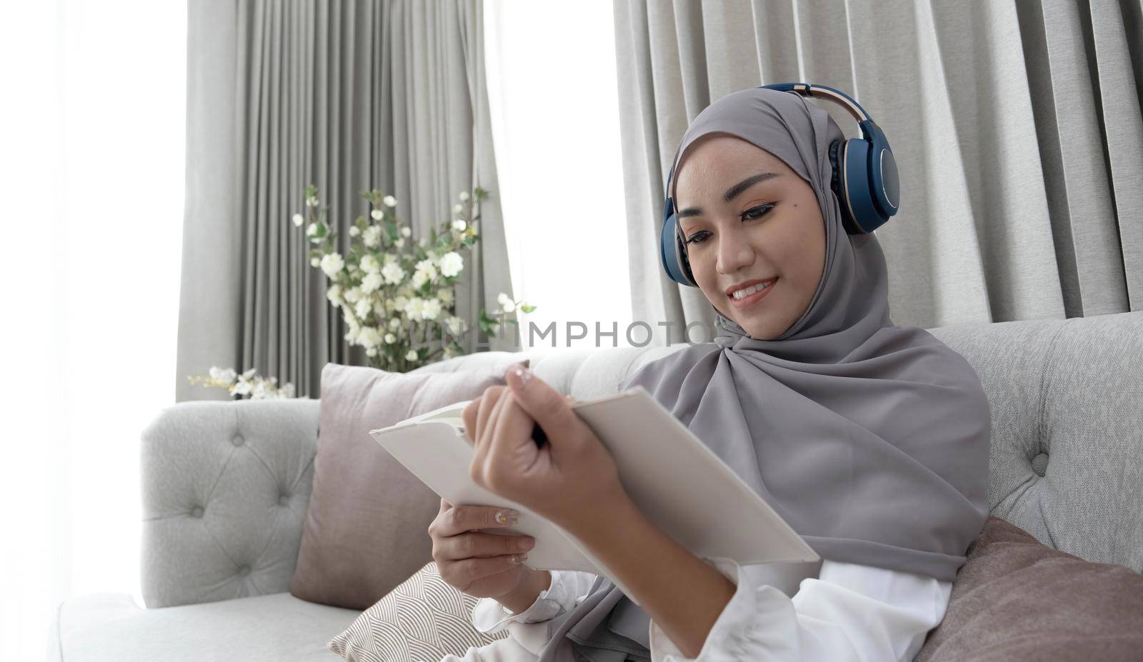 Charming young Asian Muslim woman wearing hijab and headphones, listening to music and reading a book in the living room. by wichayada