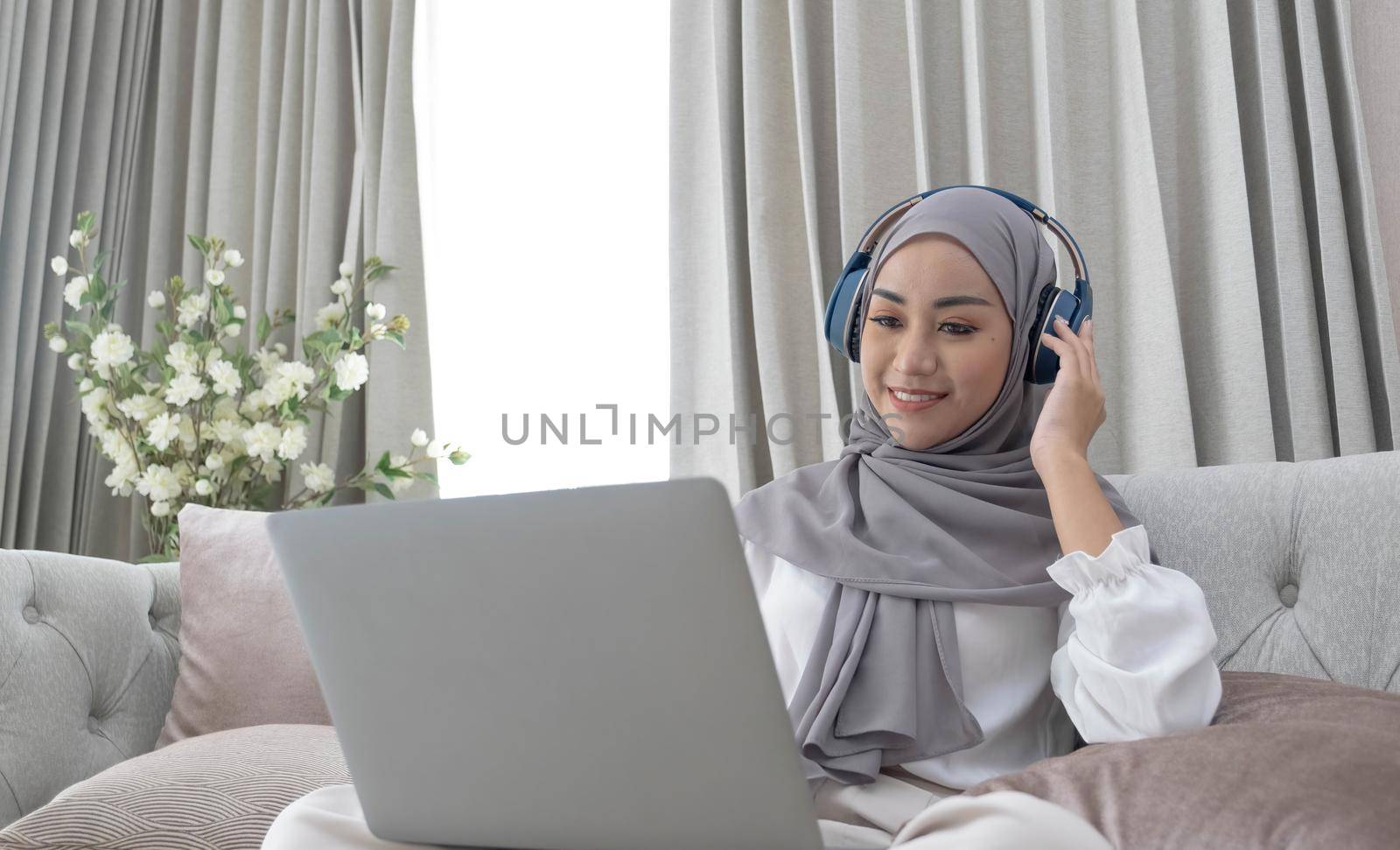 Attractive young Asian Muslim woman wearing hijab and headphones, watching movie on laptop computer on sofa. by wichayada