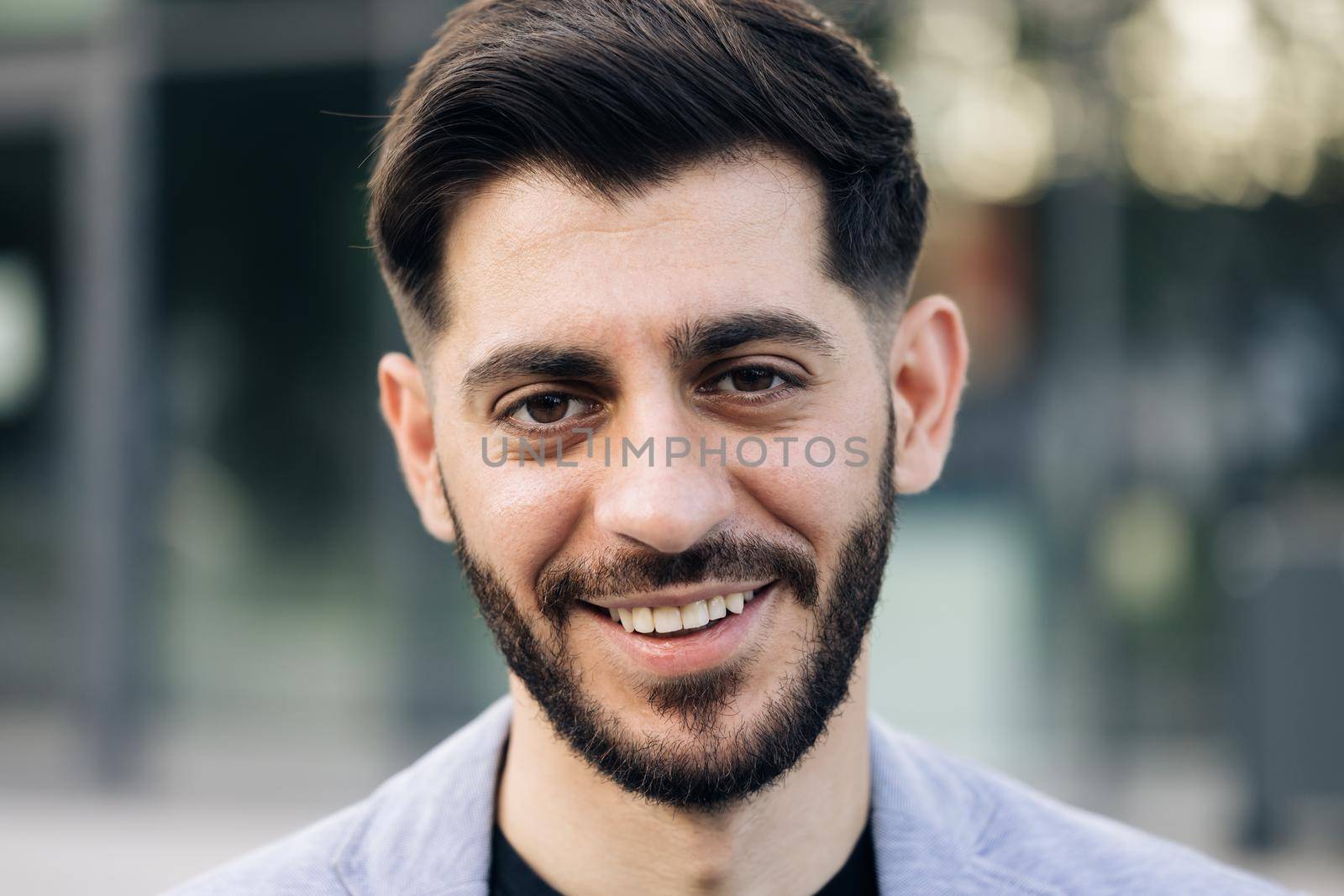 Face portrait of attractive joyful young caucasian mixed race man in business formal suit smiling and posing near modern buildings outside. Businessman. Ethnicity by uflypro