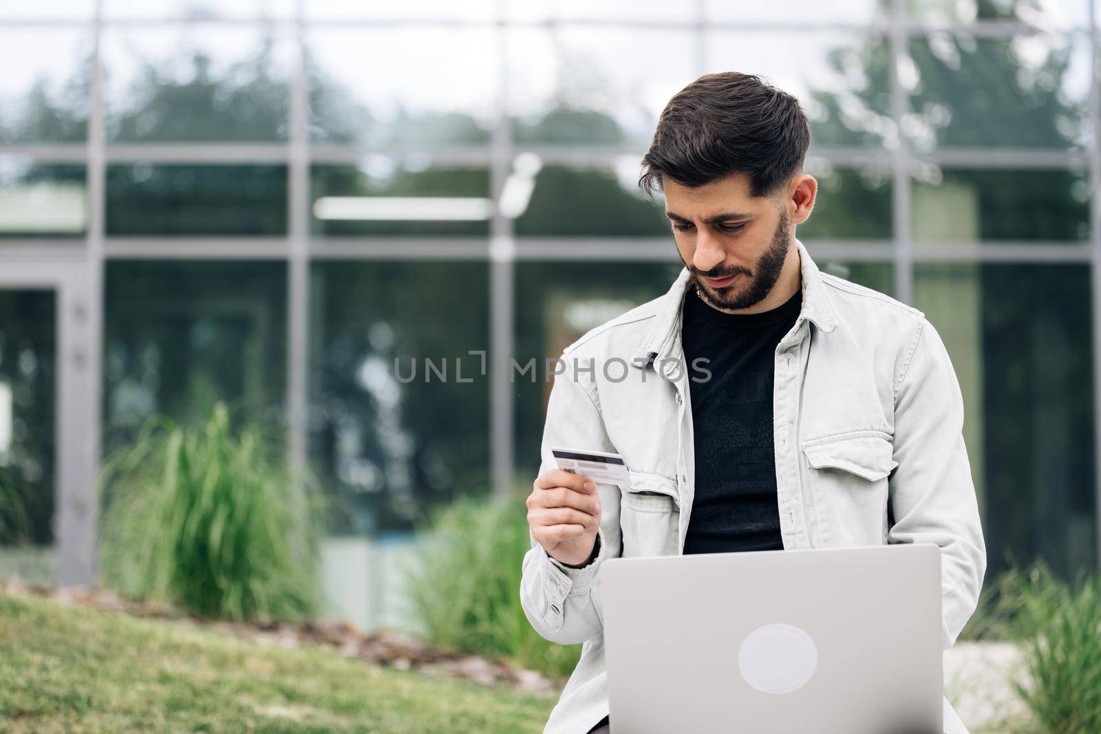 Man customer using computer e-banking service making secure online payment holding credit card in hand. Male buyer paying on website via e commerce service sitting on bench near modern office.