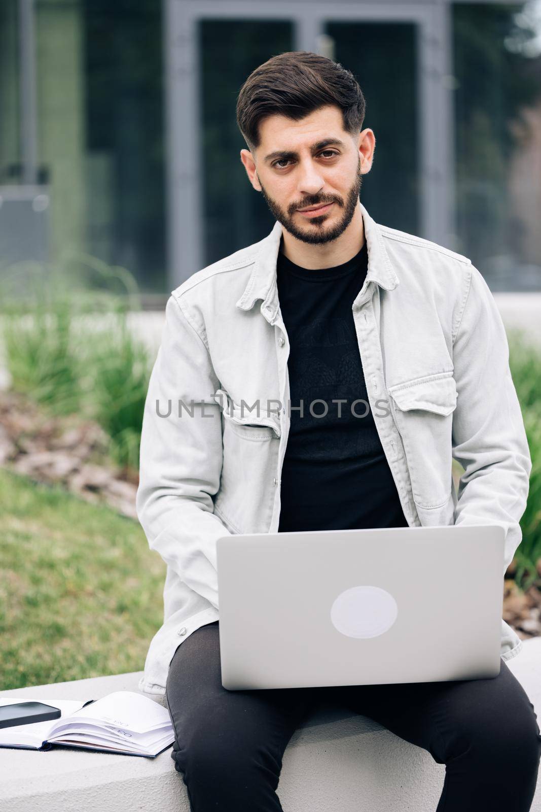 Appealing armenian bearded man typing on a laptop keyboard while sitting on the bench with a city view at day time by uflypro