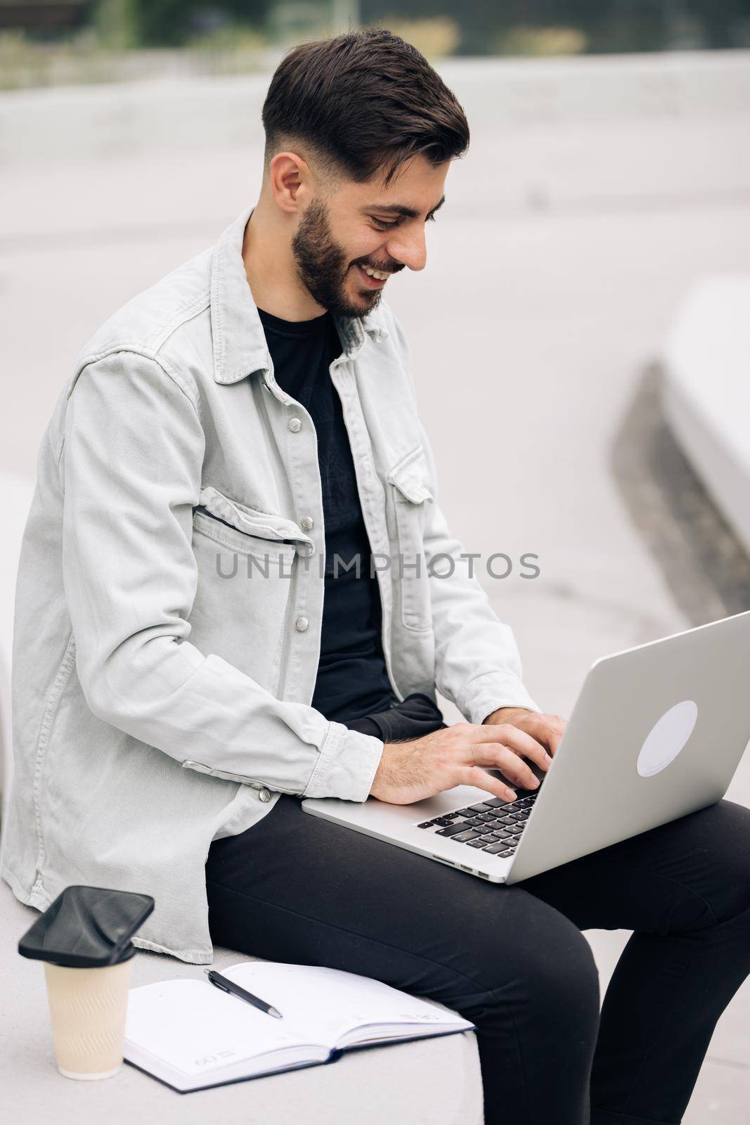 Smiling bearded businessman working on laptop computer near modern office. Male professional typing on laptop keyboard at workplace. Portrait of positive business man looking at laptop screen outdoors