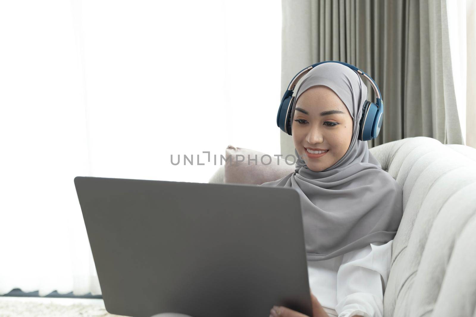 Charming young Asian Muslim woman wearing hijab sits on a comfy sofa, using a laptop computer and listening to music on her headphones. by wichayada