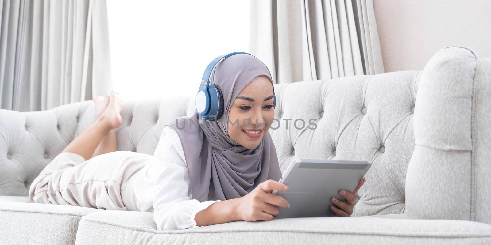 Relaxed and happy asian Muslim woman with hijab laying on sofa, listening music on headphones and using digital tablet..