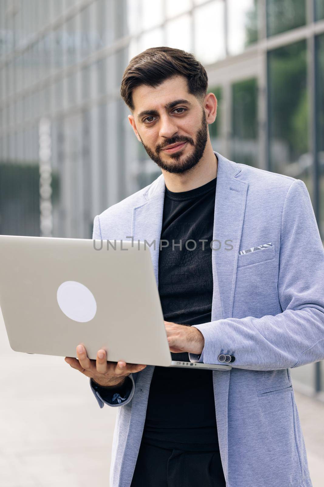 Young successful bearded businessman freelancer is staing near modern office looking and smiling at the camera. Man is working on his laptop.
