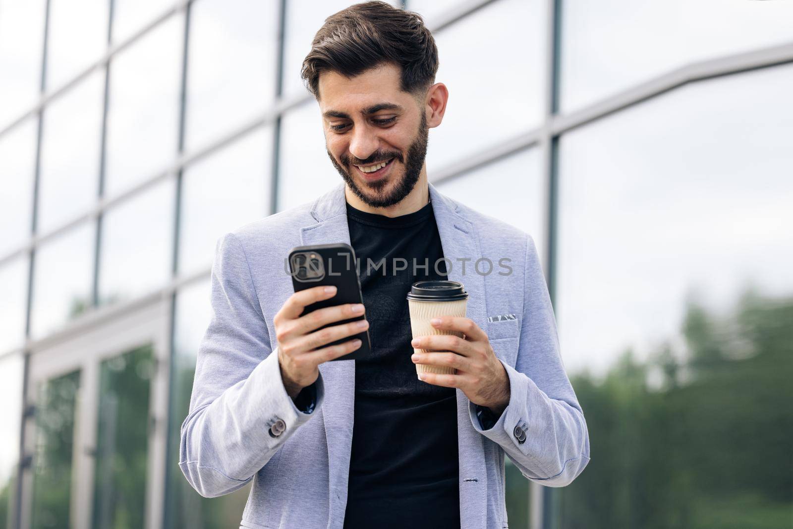 Caucasian armenian man using mobile phone holding coffee cup in modern city district. Positive business guy browsing financial news smiling standing outdoor. Successful adult man by uflypro