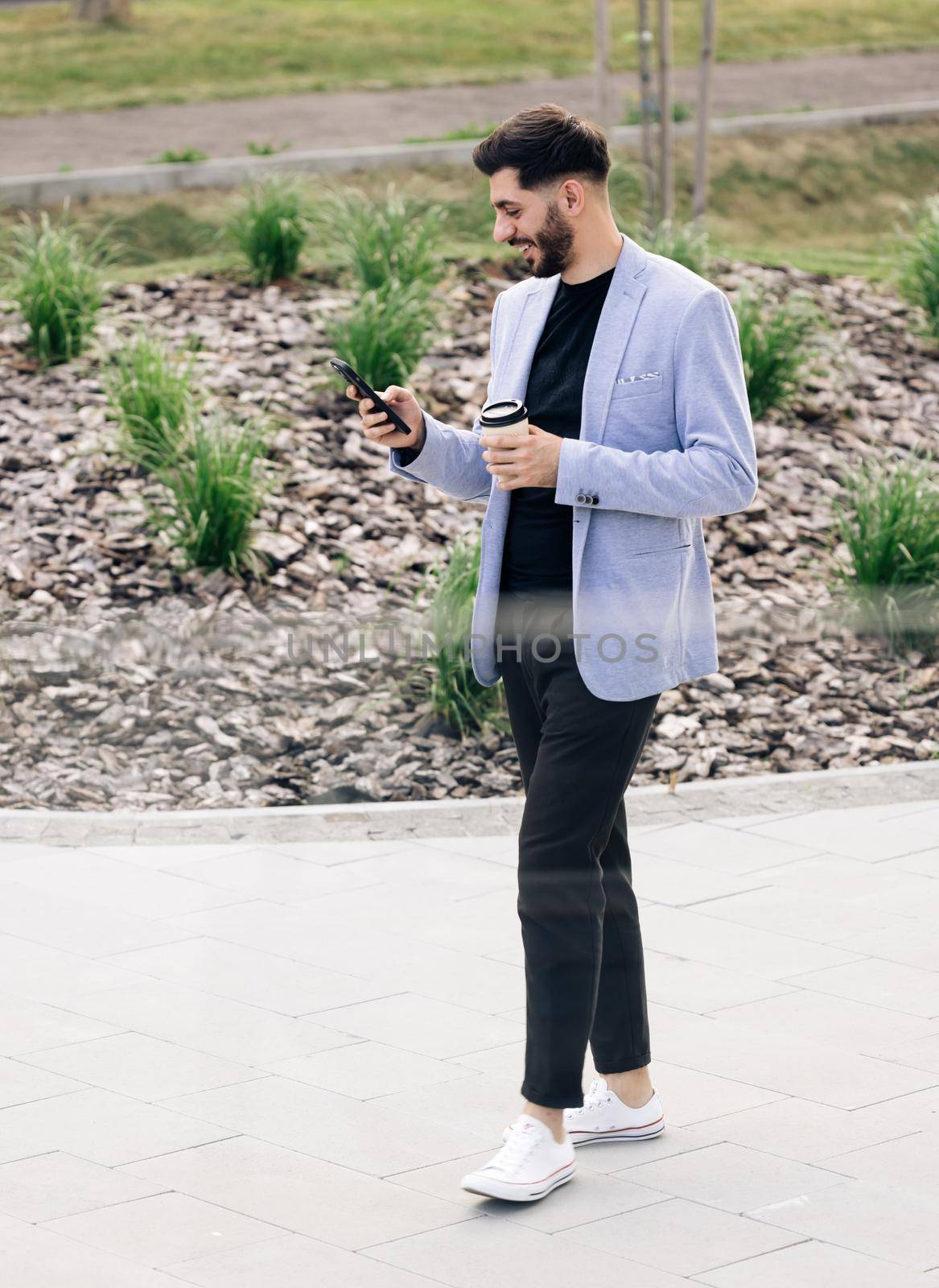 Confident armenian man using mobile phone holding coffee cup in modern city district. Positive business guy browsing financial news smiling standing outdoor. Successful adult man by uflypro