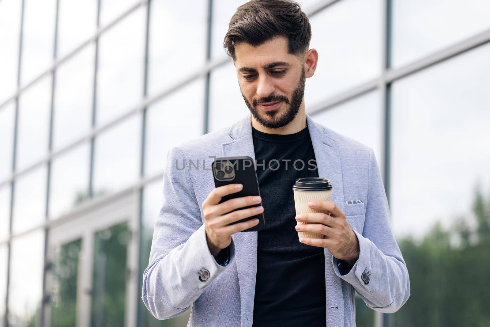 Armenian man using mobile phone holding coffee cup in modern city district. Positive business guy browsing financial news smiling standing outdoor. Successful adult man.