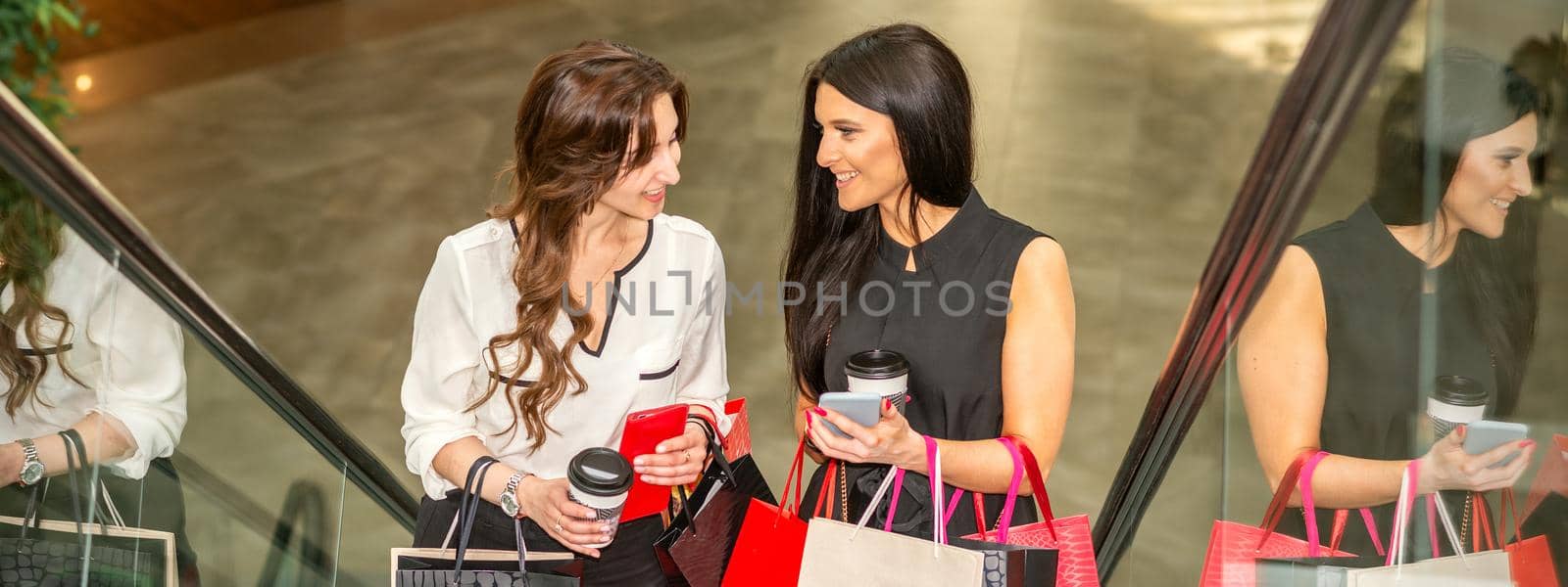 Two girlfriends going up the escalator with shopping bags and talking in the shopping mall