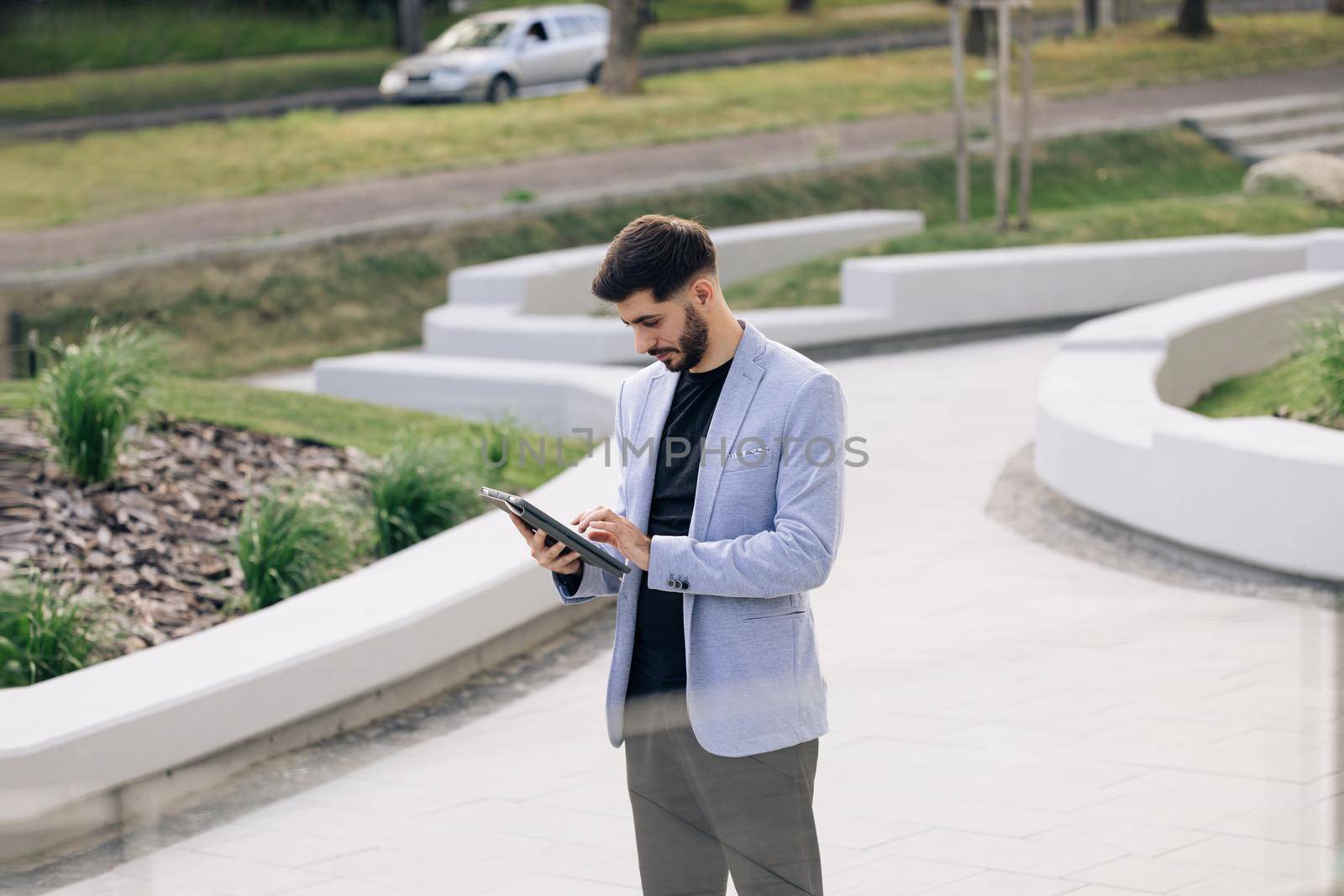 Businessman holding tablet in hands using business apps on tablet computer. Happy face looking tablet screen outdoors. Elegant male chatting online with digital device outside. by uflypro