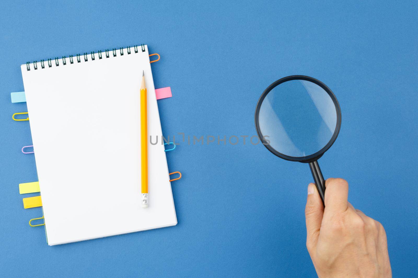 Spiral notebook with bookmarks from paper clips, note sheets, pencil, magnifying glass in hand on blue isolated background. Office concept. Top view.