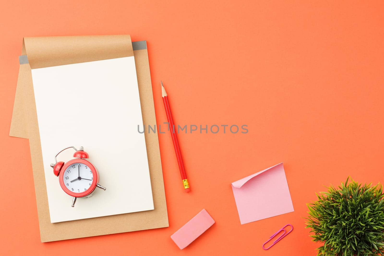 Craft notebook with white sheets, red alarm clock and pencil, rubber band, paper clip and note sheet on orange isolated background top view.