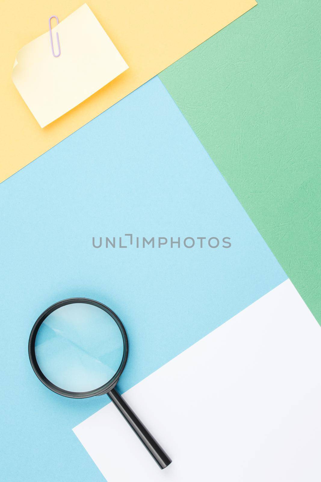 Colored paper background of geometric shapes with magnifying glass, note paper and paper clip. Office composition. Flat lay