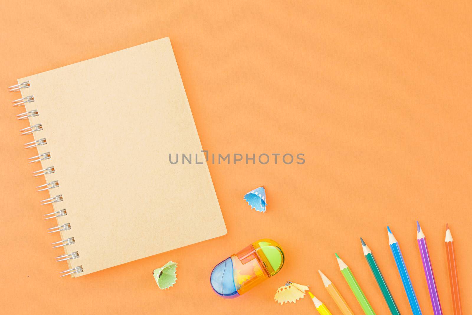 Spiral craft notebook with colored pencils sharpener and shavings on an orange background. Top view, copy space.