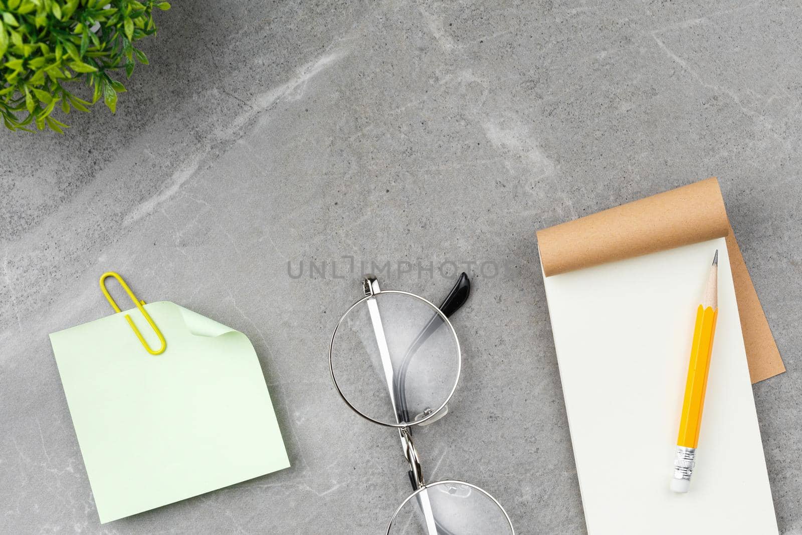 Open notepad, pencil, glasses, note paper and a flower in a pot on a gray marble background. Top view.