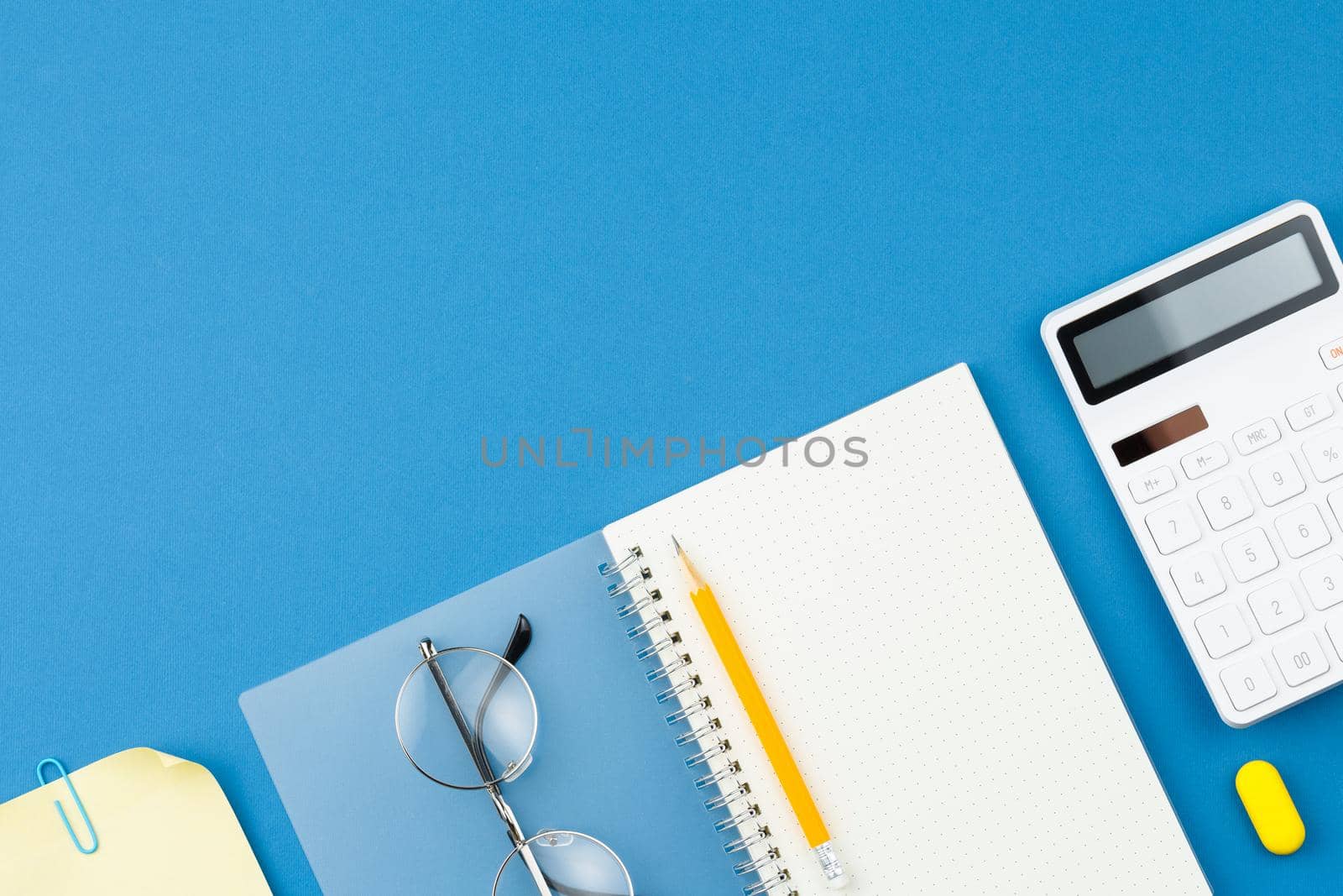 Open spiral notebook with pencil, calculator, note sheet, glasses, paper clip and rubber band on blue isolated background. Office concept. Top view.