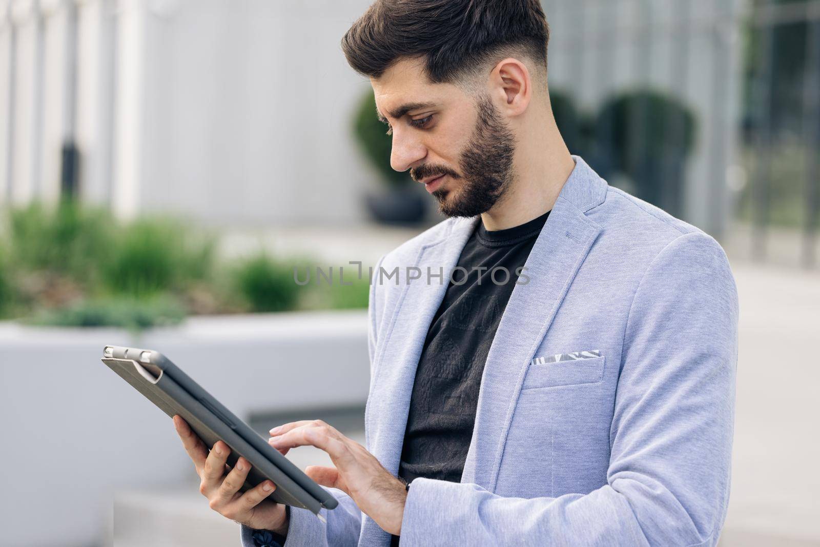 Elegant male chatting online with digital device outside. Businessman holding tablet in hands using business apps on tablet computer. Happy face looking tablet screen outdoors by uflypro