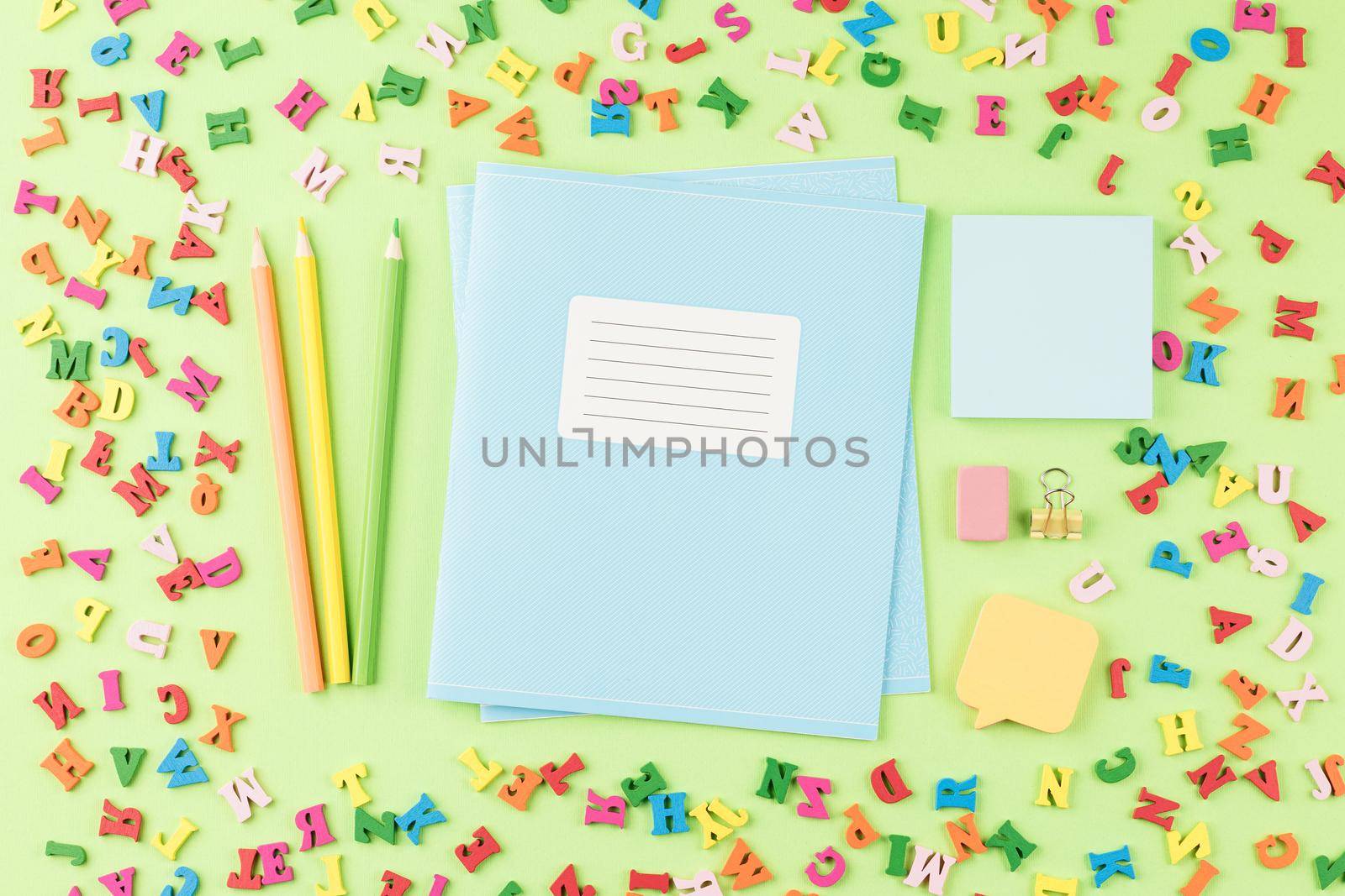 School notebook with colored pencils on a background of multicolored wooden letters. Flat lay. Back to school concept. Paper stickers on a green table. Top view.