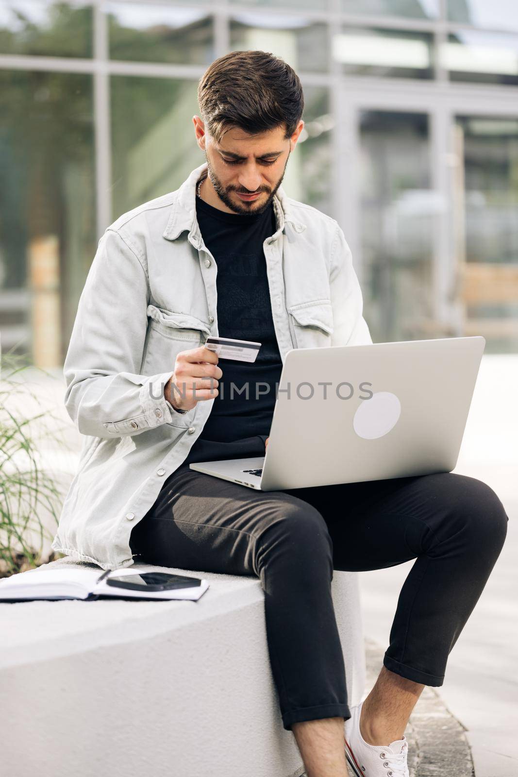 Male buyer paying on website via e commerce service sitting on bench near modern office. Man customer using computer e-banking service making secure online payment holding credit card in hand.
