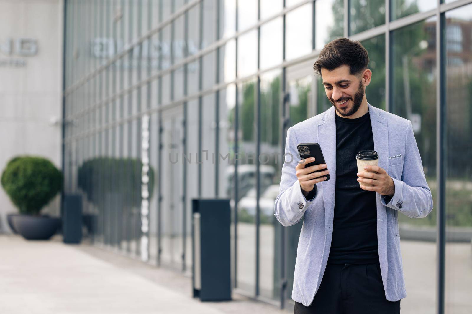 Confident caucasian armenian man using mobile phone holding coffee cup in modern city district. Positive business guy browsing financial news smiling standing outdoor. Successful adult man.
