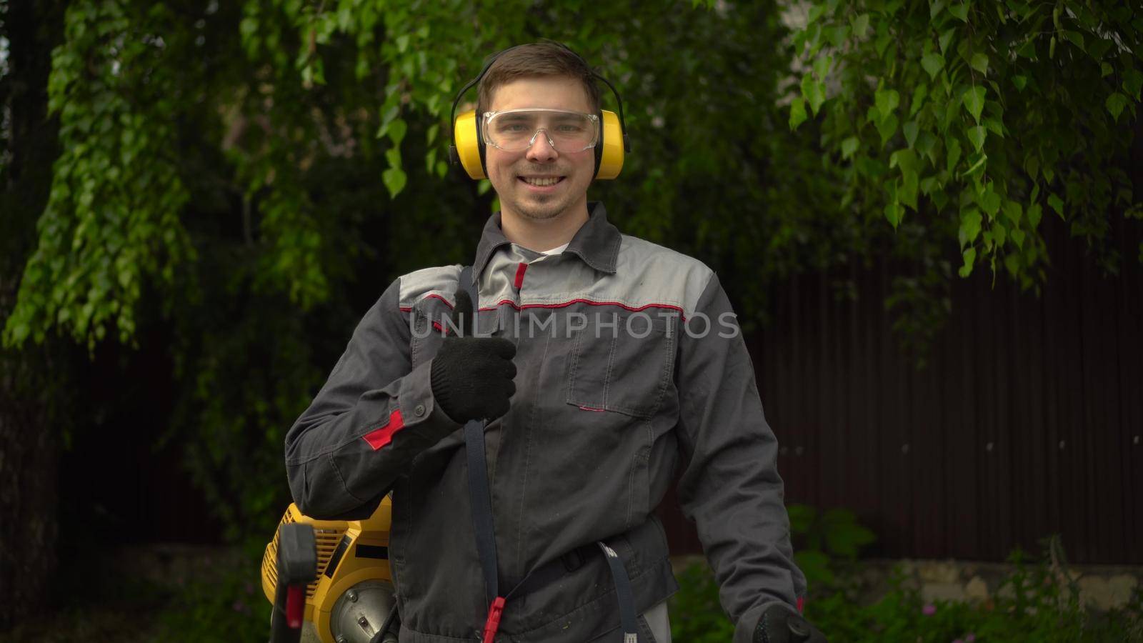 A young man from the special services mows the lawn with a petrol trimmer. A man in glasses and headphones looks at the camera, smiles and shows a thumbs up. 4k