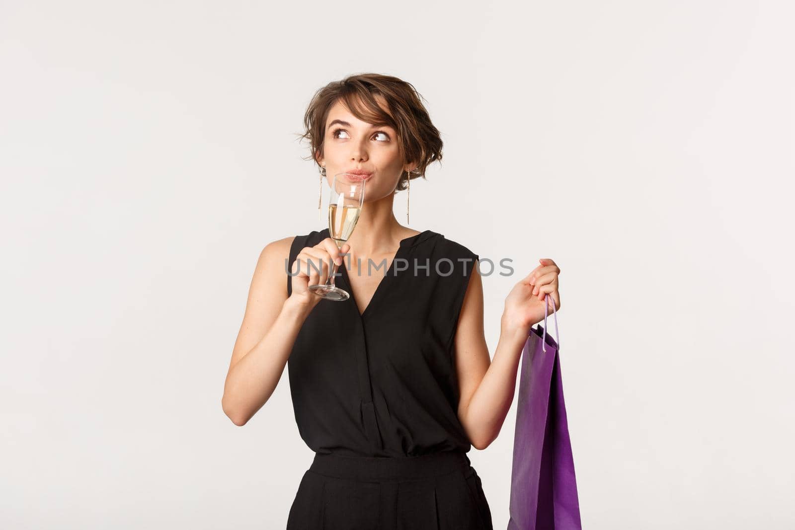 Thoughtful stylish woman drinking champagne, holding shopping bad and looking upper left corner dreamy, standing over white background.