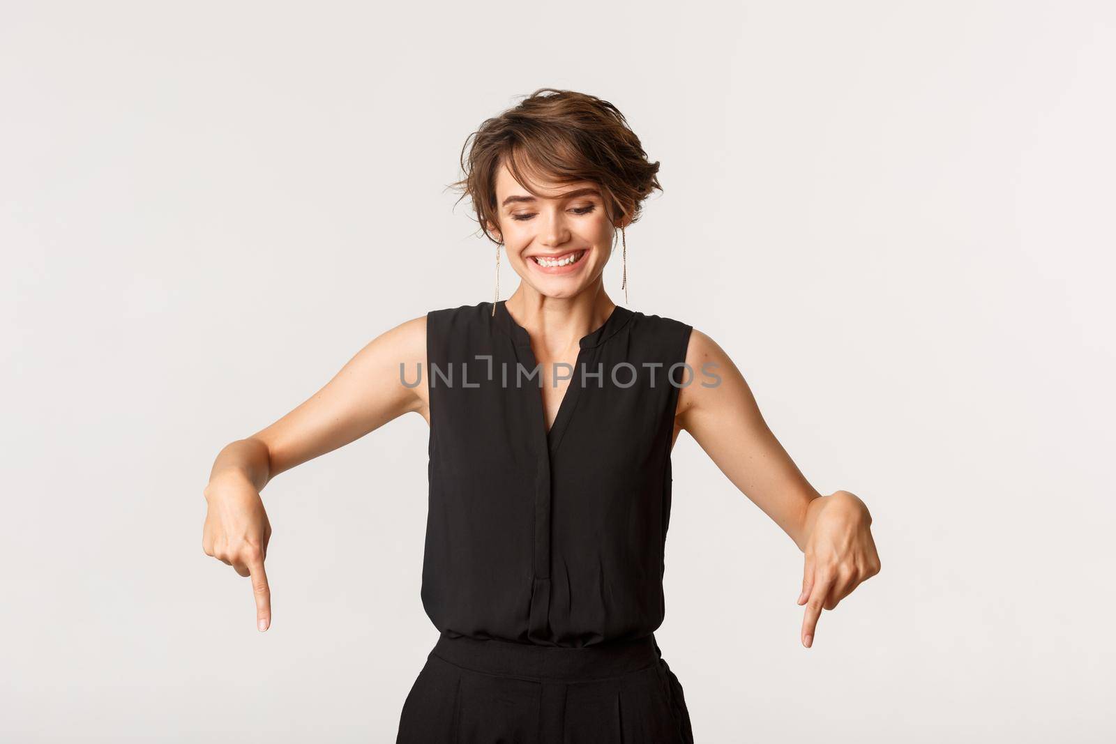 Happy adult woman in black dungarees pointing fingers down, looking downwards with pleased smile by Benzoix