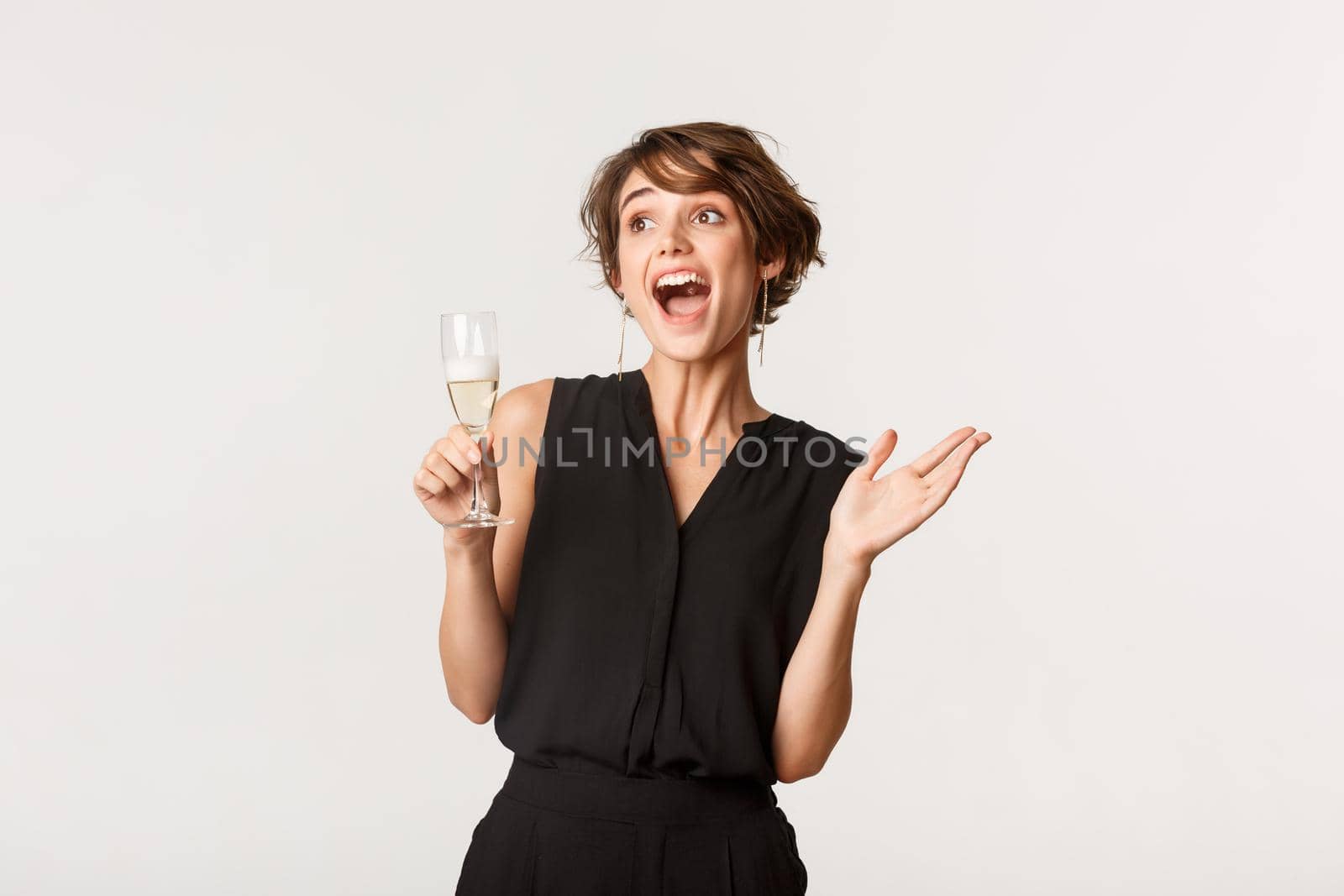 Excited party girl looking joyful upper left corner, holding glass of champagne, standing over white background.