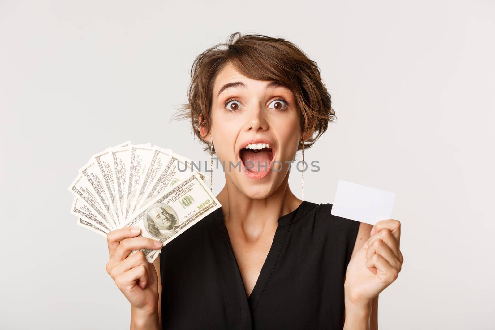 Close-up of amazed young woman open mouth fascinated, holding money and credit card, standing over white background by Benzoix