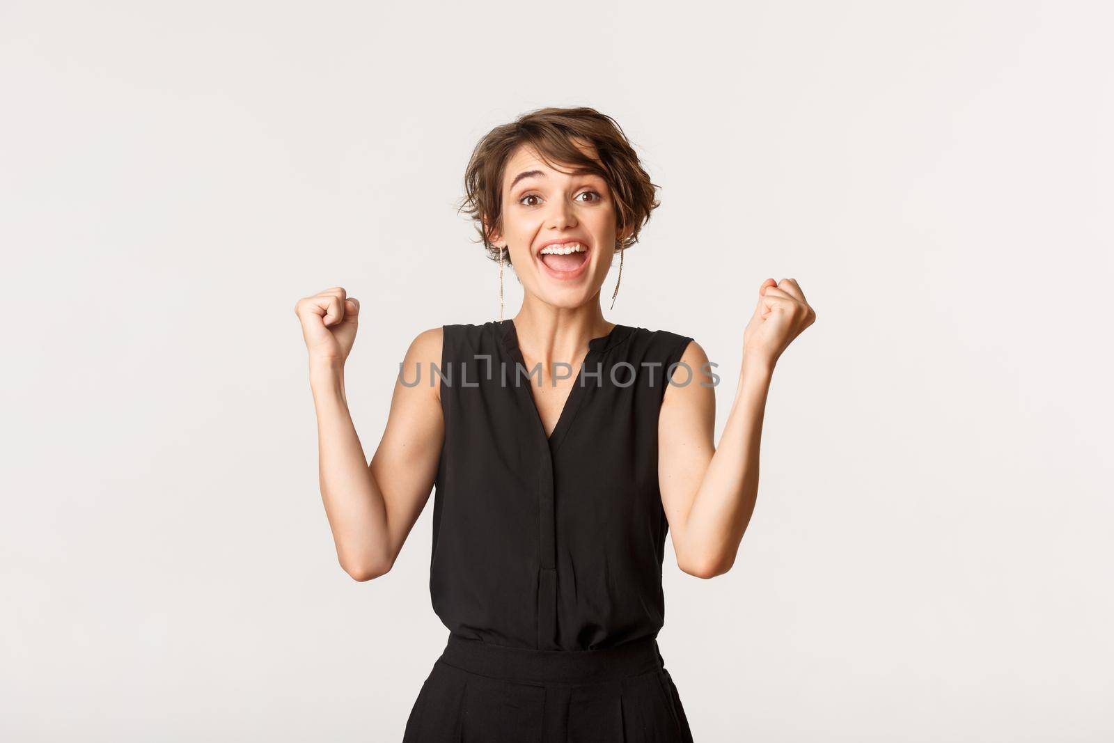 Excited successful young woman feeling lucky, smiling upbeat and fist pump, celebrating victory over white background.