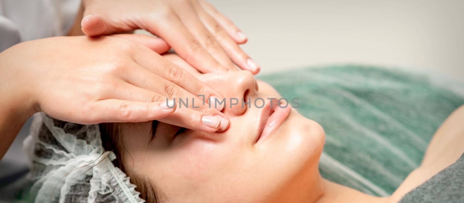 Young woman getting facial massage with closed eyes by beautician in beauty salon