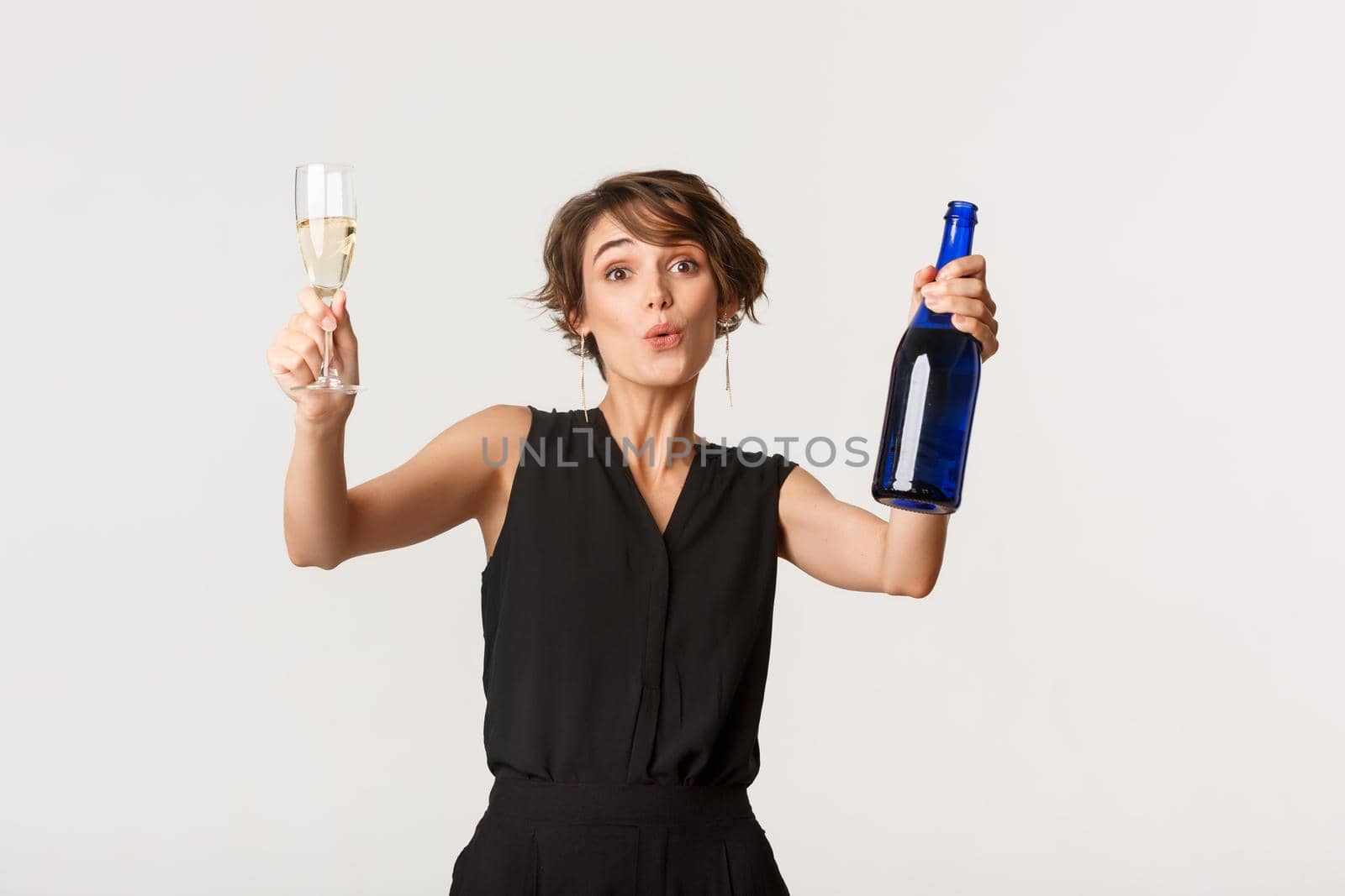 Cheerful elegant woman having fun on party, celebrating something, dancing with bottle and glass of champagne, standing over white background.