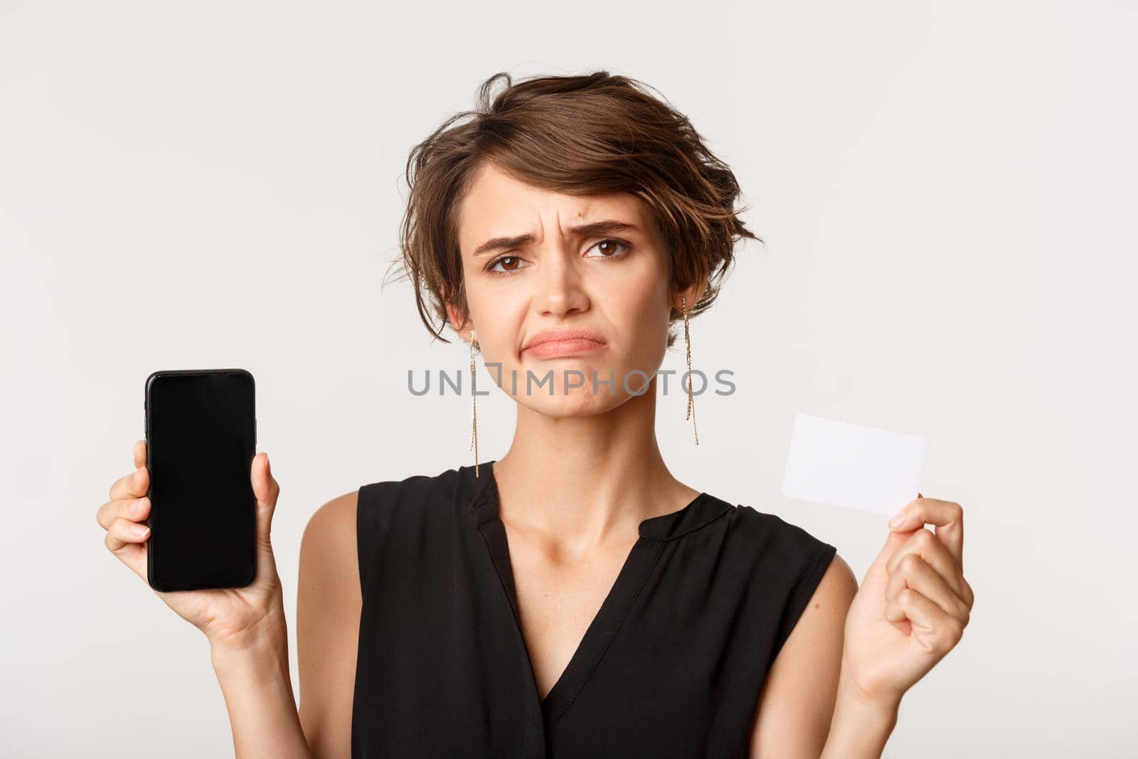 Close-up of disappointed, skeptical brunette woman smirk unamused, showing credit card and smartphone screen, standing over white background.