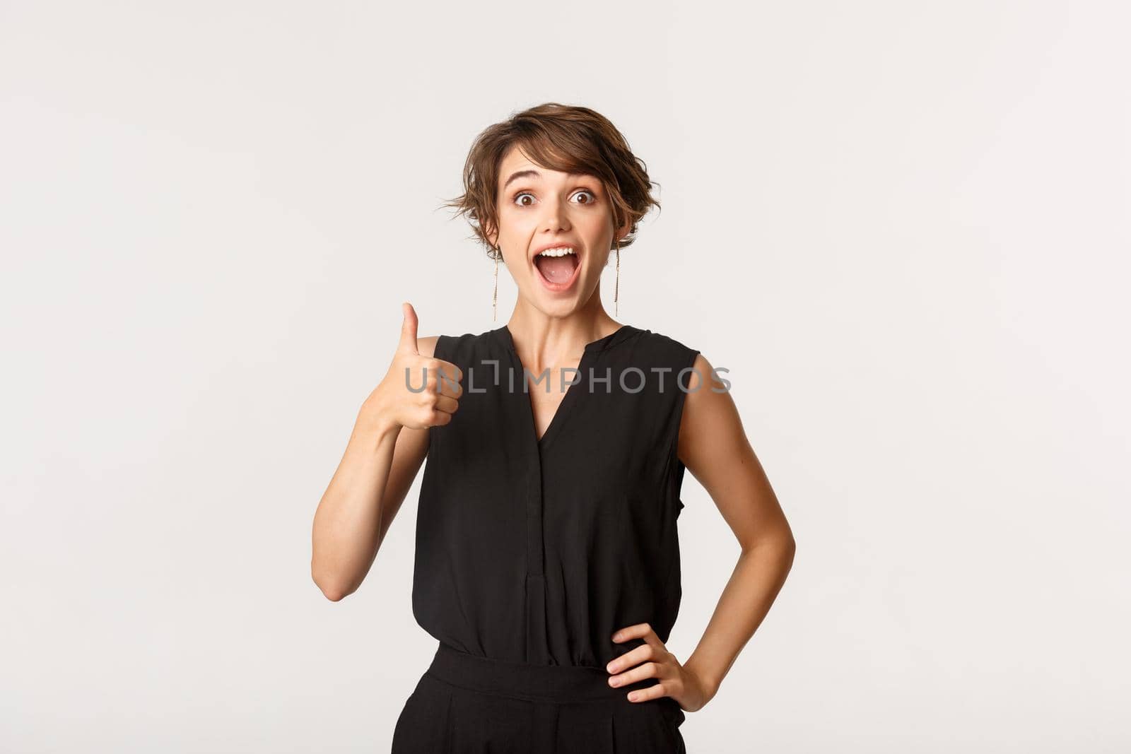 Amazed and satisfied young woman showing thumbs-up with excited face, standing white background.