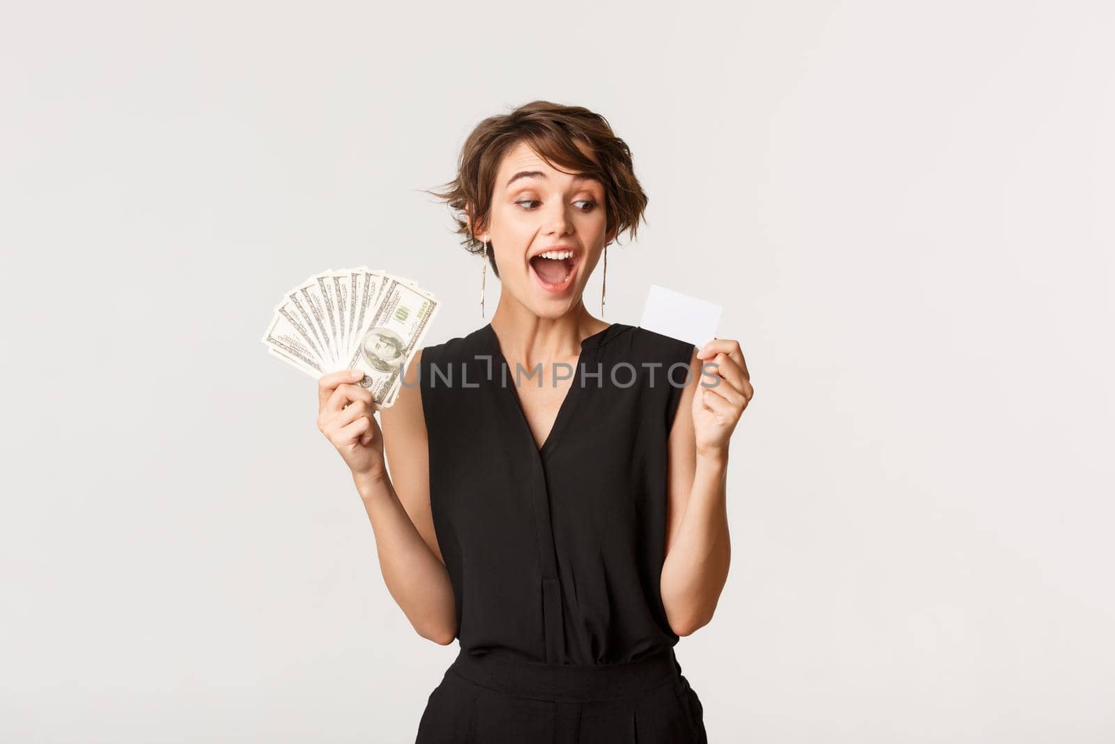 Amazed elegant woman looking at credit card, holding money, standing white background.