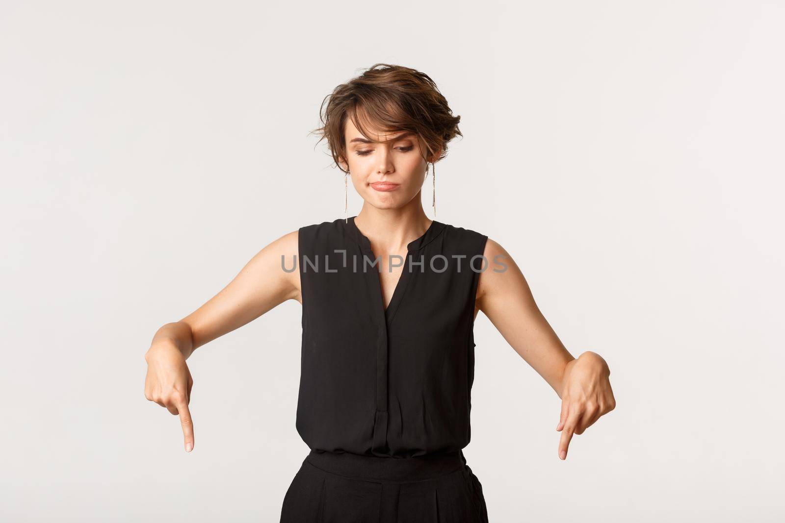 Skeptical and doubtful young caucasian woman smirk hesitant, pointing and looking down, white background.