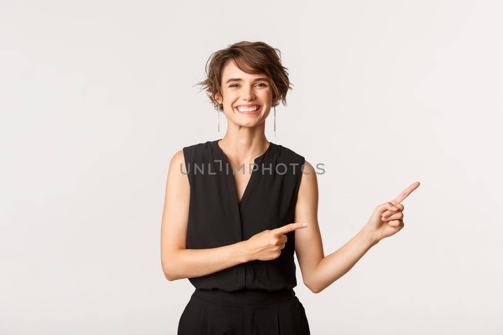 Happy gorgeous young woman in black elegant clothes pointing fingers upper right corner and smiling cheerful, showing advertisement over white background by Benzoix