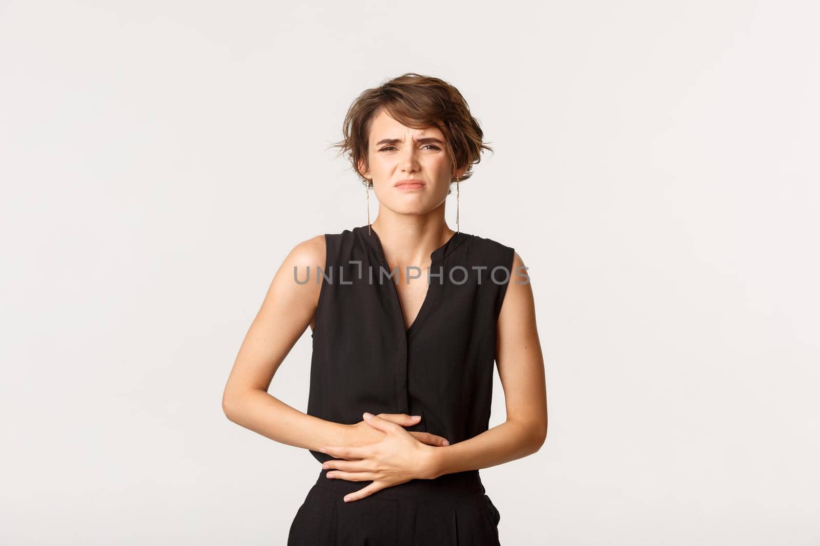 Woman in formal outfit touching belly and grimacing from pain. Girl having painful cramps or stomach ache, standing white background feeling discomfort by Benzoix