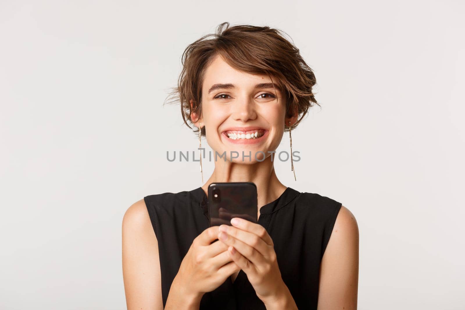 Close-up of happy woman holding smartphone, smiling at camera, standing over white background by Benzoix