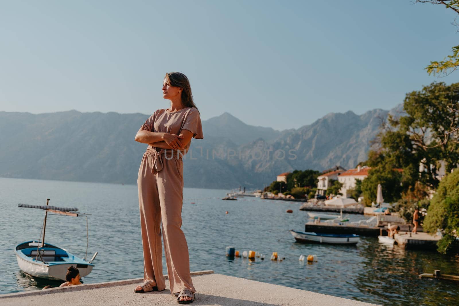 Girl Tourist Walking Through Ancient Narrow Street On A Beautiful Summer Day In MEDITERRANEAN MEDIEVAL CITY, MONTENEGRO. Young Beautiful Cheerful Woman Walking On Old Street At Tropical Town. Pretty Girl Looking At You And Smiling by Andrii_Ko