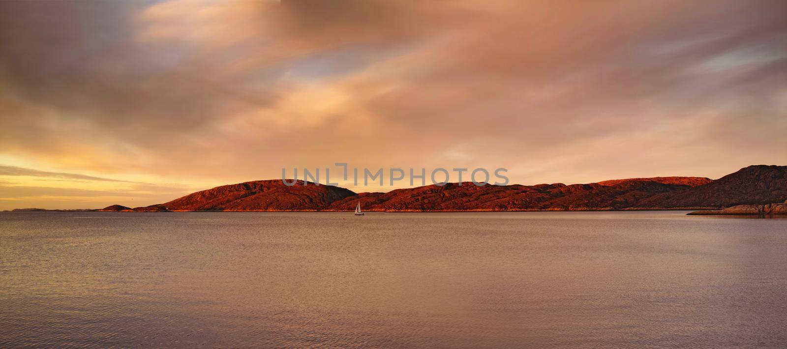 Distant sea landscape near a mountain with a small sailboat in the water. The ocean, beach, or large lake with a boat in the distance near a hill outdoors during sunset on a cloudy day.