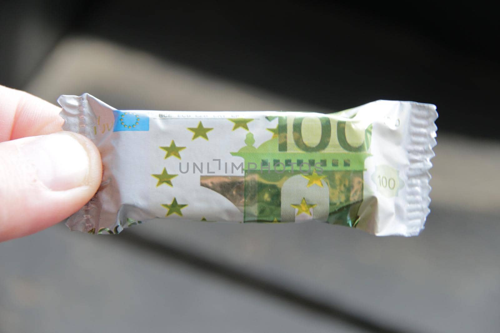 A hand holds a candy with a wrapper in the form of a one hundred euro banknote by Markgraf