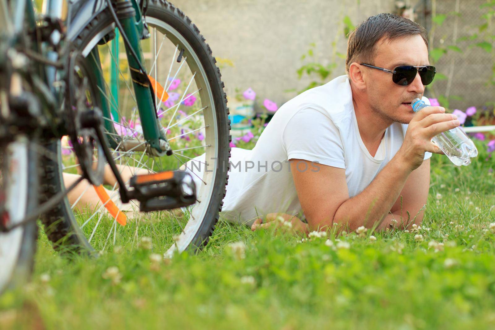 Close up view of man lying on green grass and drinking water from a bottle. Picture of male in white T-shirt and sunglasses with bottle of water lying on green grass with road bicycle beside him in the garden. Selective focus on man. Healthy active lifestyle