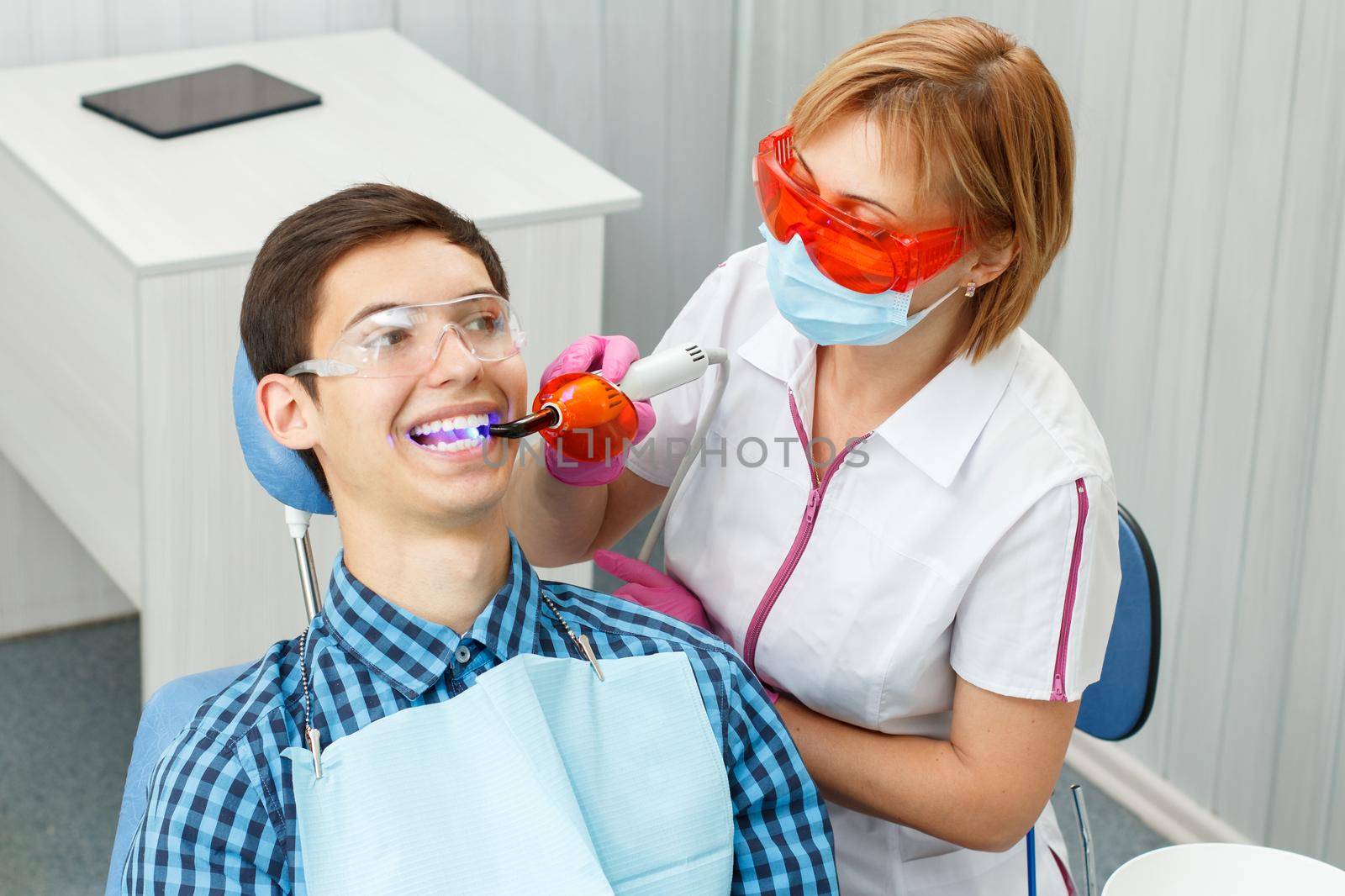Beautiful woman dentist treating a patient teeth in dental office. Doctor wearing glasses, mask, white uniform and pink gloves. Dentistry