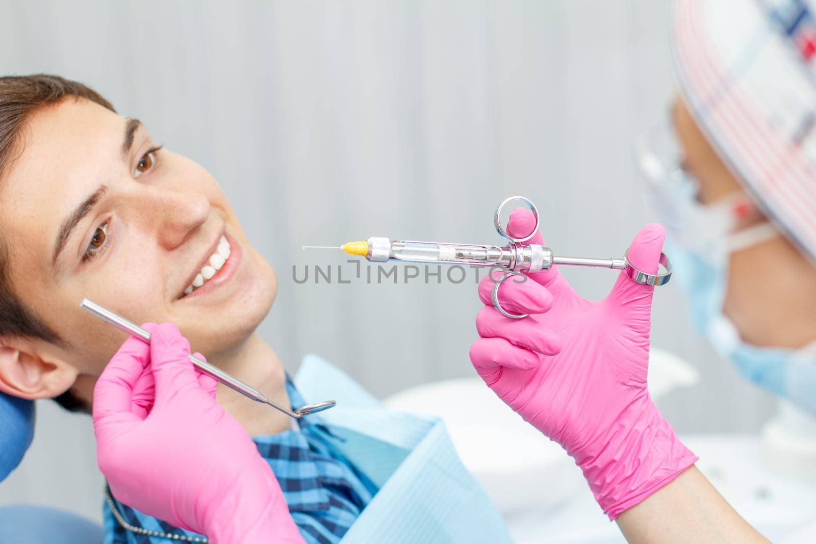 Doctor wearing pink gloves is holding syringe with anesthetic and mirror with smiling patient on the background. Selective focus on hand with syringe. Application of anesthesia in dentistry